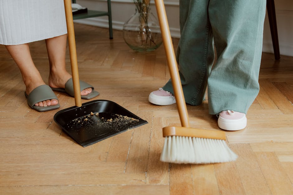 Close-up view of two individuals performing surface cleaning in a domestic setting, focusing on their feet and the cleaning tools. One person is wearing grey slide slippers and pale beige pants, standing near a black dustpan with crumbs, while the other person is dressed in light blue jeans and white sneakers, holding a broom with a wooden handle. The wooden floor, made of light-colored hardwood, appears clean and polished, with minimal dust or debris visible. In the background, part of a wooden table leg and a glass vase with decorative plants can be seen, under natural lighting that highlights the fresh and tidy environment. This scene illustrates routine domestic cleaning, emphasizing hygiene and surface maintenance, consistent with Bromley Carpet Cleaning's professional cleaning services in Bromley.