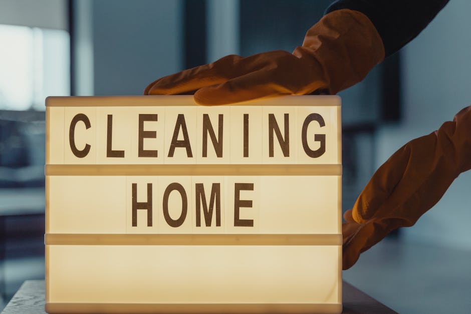 A person wearing orange cleaning gloves is adjusting a brightly lit lightbox sign that displays the words 'CLEANING HOME' in bold black letters on a white background. The scene appears to be set indoors with a modern, clean environment, featuring smooth surfaces and subtle lighting. The image emphasizes the concept of home surface cleaning and sanitisation, relevant to residential and commercial cleaning services offered by Bromley Carpet Cleaning, with a focus on maintaining hygiene and deep cleaning to avoid hidden charges in Bromley homes.