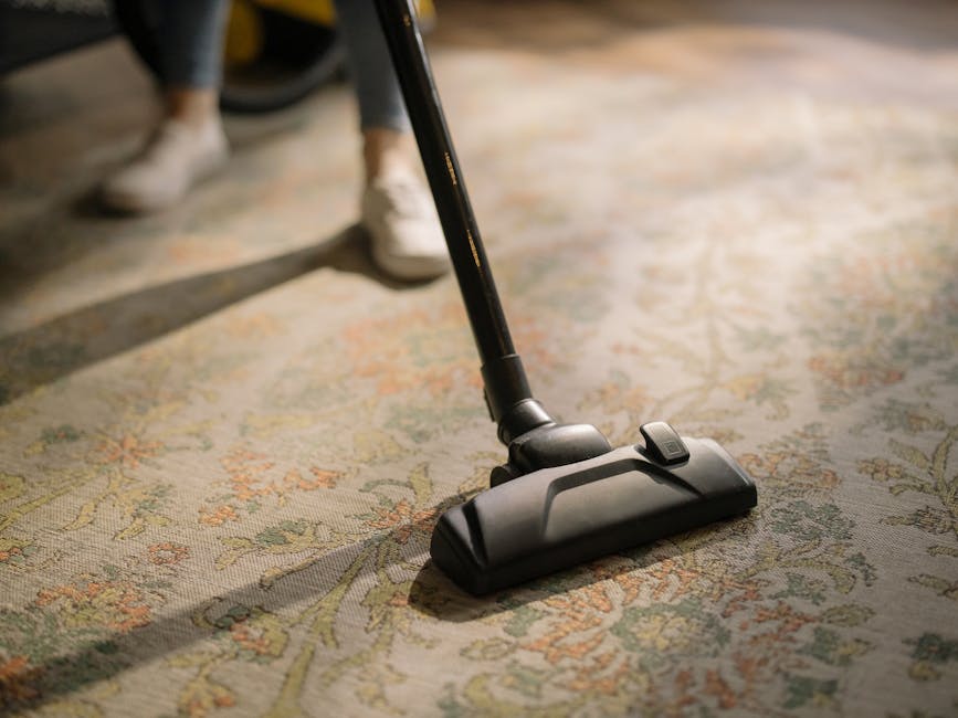 Close-up image of a vacuum cleaner head attached to a black hose, actively cleaning an intricate, multicolored patterned rug with floral motifs in a room. The floor beneath appears to be part of a living or common area with soft, diffused lighting illuminating the cleaned surface. The scene suggests a thorough surface cleaning process by Bromley Carpet Cleaning, emphasizing professional deep cleaning and sanitisation of domestic carpets.