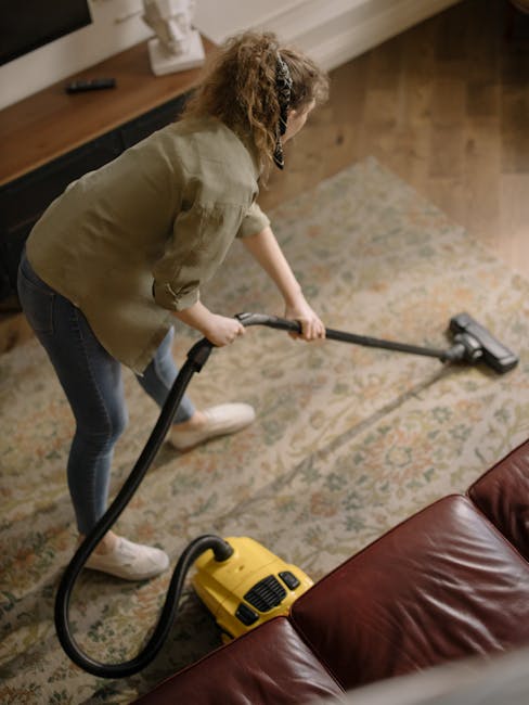 A woman with curly hair, wearing a beige jacket, blue jeans, and white shoes, is engaged in surface cleaning using a yellow vacuum cleaner with a handheld attachment, on a patterned area rug in a living room. The room features wooden flooring, a brown leather sofa, and a sideboard against the wall with decorative items, including a white sculpture. Bright, natural lighting illuminates the space, highlighting the cleanliness and neat arrangement. Bromley Carpet Cleaning offers professional domestic cleaning services focused on deep cleaning and surface sanitisation, as seen in this scene.