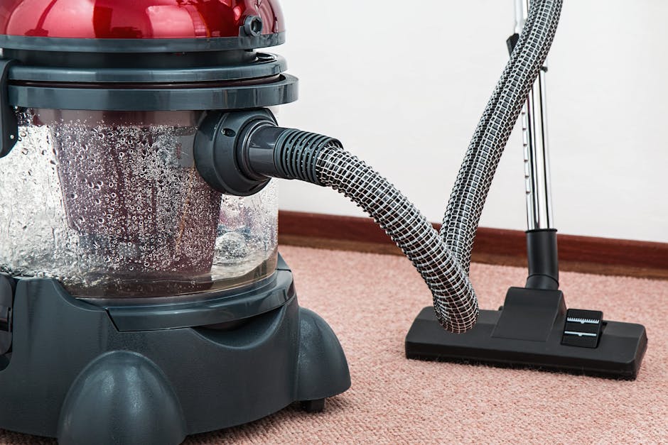 A close-up view of a vacuum cleaner with a transparent dust container, showing dust and debris inside. The vacuum has a black and red body with a flexible, patterned hose attached to a flat, rectangular cleaning head that is resting on a pink carpeted floor. The background features a white wall and a brown wooden skirting board. The scene highlights the process of surface cleaning and deep cleaning in a domestic setting, with the vacuum cleaner used for removing dust and dirt for hygiene and maintenance purposes. The image is used to illustrate cleaning services offered by Bromley Carpet Cleaning, as seen on the Bromley South Station Carpet Cleaning Guide page.