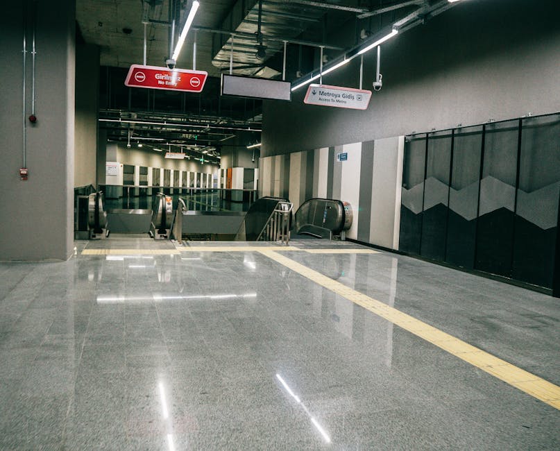 The image shows the interior of Bromley South Station with a polished, shiny granite floor reflecting overhead lighting. The floor is smooth and well-maintained, with a distinct tactile paving strip running through the middle to aid visually impaired travelers. In the background, there are two escalators descending to the platform area, with user-access control barriers on either side. The station walls feature modern geometric paneling in black and gray tones, creating a clean and contemporary appearance. Suspended above are illuminated signage panels indicating directions, including signs for 'Girliyiz No Entry' and 'Metroy Gidiş,' providing clear navigation. The lighting is bright and evenly distributed, emphasizing the cleanliness and tidiness of the space. Bromley Carpet Cleaning is mentioned as part of the context, but the focus remains on the pristine surface and overall hygiene of the station environment, highlighting aspects of surface cleaning and maintenance in a public transportation hub.