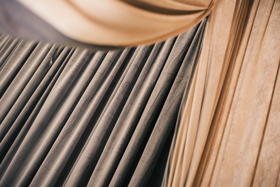 Close-up view of a set of grey curtains made of velvety fabric hanging along a wooden curtain rail, with a portion of light-colored wooden flooring visible underneath. The curtains are neatly pleated and appear clean without any visible stains or dust, suggestive of surface cleaning or regular maintenance. The lighting is soft, highlighting the texture of the fabric and the smoothness of the wooden surfaces. The image exemplifies the kind of domestic or commercial fabric surface cleaning that Bromley Carpet Cleaning offers as part of their deep cleaning and sanitisation services for maintaining hygiene and appearance in residential spaces.