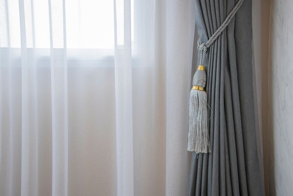 A close-up image of a window with sheer white curtains behind, allowing natural light to softly illuminate the room. To the right, a heavy grey curtain is pulled to the side and secured with a decorative grey and white tassel tieback that features gold accents. The curtains are made of fabric with a smooth, matte finish, and the window frame is not visible. The room appears clean and well-maintained, with a neutral color palette that emphasizes the clarity and freshness of the window area. The setting suggests an environment where surface cleaning and window maintenance are prioritized, consistent with services offered by Bromley Carpet Cleaning in the context of domestic cleaning for optimal hygiene and appearance.