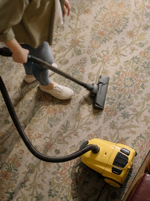 A person is performing surface cleaning on a patterned area rug using a yellow vacuum cleaner with a black hose attachment, in a room with warm lighting. The individual is wearing beige sneakers, blue jeans, and a beige jacket, standing on the rug to vacuum the intricate floral and vine design with muted colors. The scene shows a close-up view of the cleaning process, focusing on the vacuum device and the textured surface of the carpet, emphasizing hygienic maintenance in a domestic setting. This image underscores professional and routine deep cleaning techniques promoted by Bromley Carpet Cleaning for maintaining home hygiene, as highlighted in the Hayes Lane Carpet Cleaning Tips page for Bromley homes.
