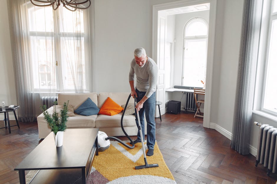A middle-aged man with gray hair and glasses is vacuuming a light wooden parquet floor in a bright living room. The room features large windows with sheer white curtains, allowing natural light to illuminate the space. A white sofa with blue and orange cushions sits against the wall, with a small black side table holding a glass of water nearby. A yellow and white patterned rug covers part of the floor, and a green potted plant is placed on a dark wooden coffee table. The man is dressed in a light gray sweater and dark pants, using a modern vacuum cleaner with a black hose and silver body. The room appears clean and well-maintained, with a minimalist decor style, and includes a small desk and a folding chair near the window as well as a radiator beneath the windows. Bromley Carpet Cleaning emphasizes professional surface cleaning, deep cleaning, and sanitisation for residential spaces like this one.