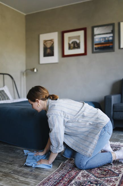 A person is performing surface cleaning on a patterned area rug using a yellow vacuum cleaner with a black hose attachment, in a room with warm lighting. The individual is wearing beige sneakers, blue jeans, and a beige jacket, standing on the rug to vacuum the intricate floral and vine design with muted colors. The scene shows a close-up view of the cleaning process, focusing on the vacuum device and the textured surface of the carpet, emphasizing hygienic maintenance in a domestic setting. This image underscores professional and routine deep cleaning techniques promoted by Bromley Carpet Cleaning for maintaining home hygiene, as highlighted in the Hayes Lane Carpet Cleaning Tips page for Bromley homes.